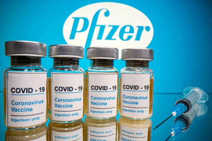 quatro frascos contendo a vacina contra a covid-19 da pfizer, em um fundo azul. Em seguida, uma seringa deitada sobre uma superfície de vidro