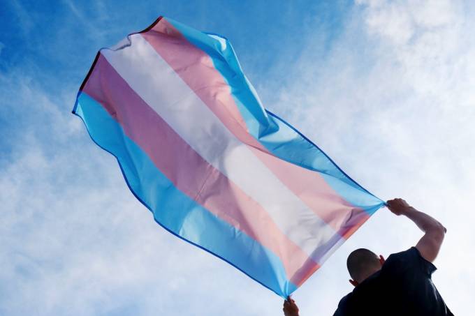 Um homem trans hasteia a bandeira do movimento trans sob o céu, capturado de baixo para cima.