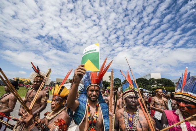 grupo de indígenas empunhando a constituição. ao fundo, um céu azul cheio de nuvens